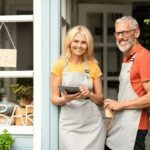 Two smiling mature adults wearing aprons are standing in front of a small business in Charlotte, NC, with an 'open' sign.