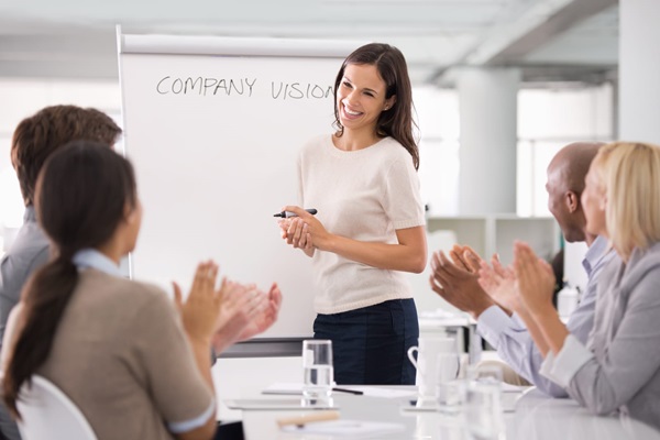 A woman presenting "company vision" on a whiteboard to a group of colleagues who are applauding.